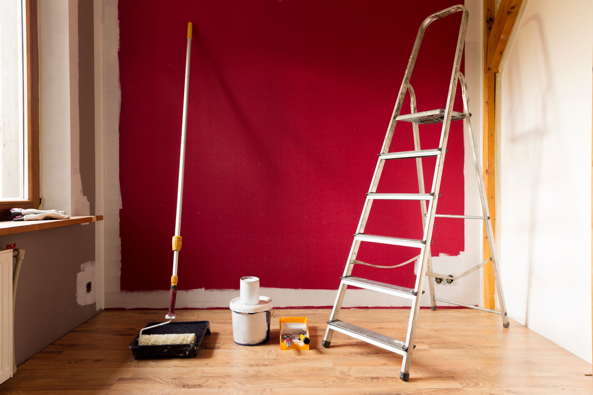 Pièce en rénovation avec un mur rouge, une échelle, du matériel de peinture et des outils sur un sol en bois.