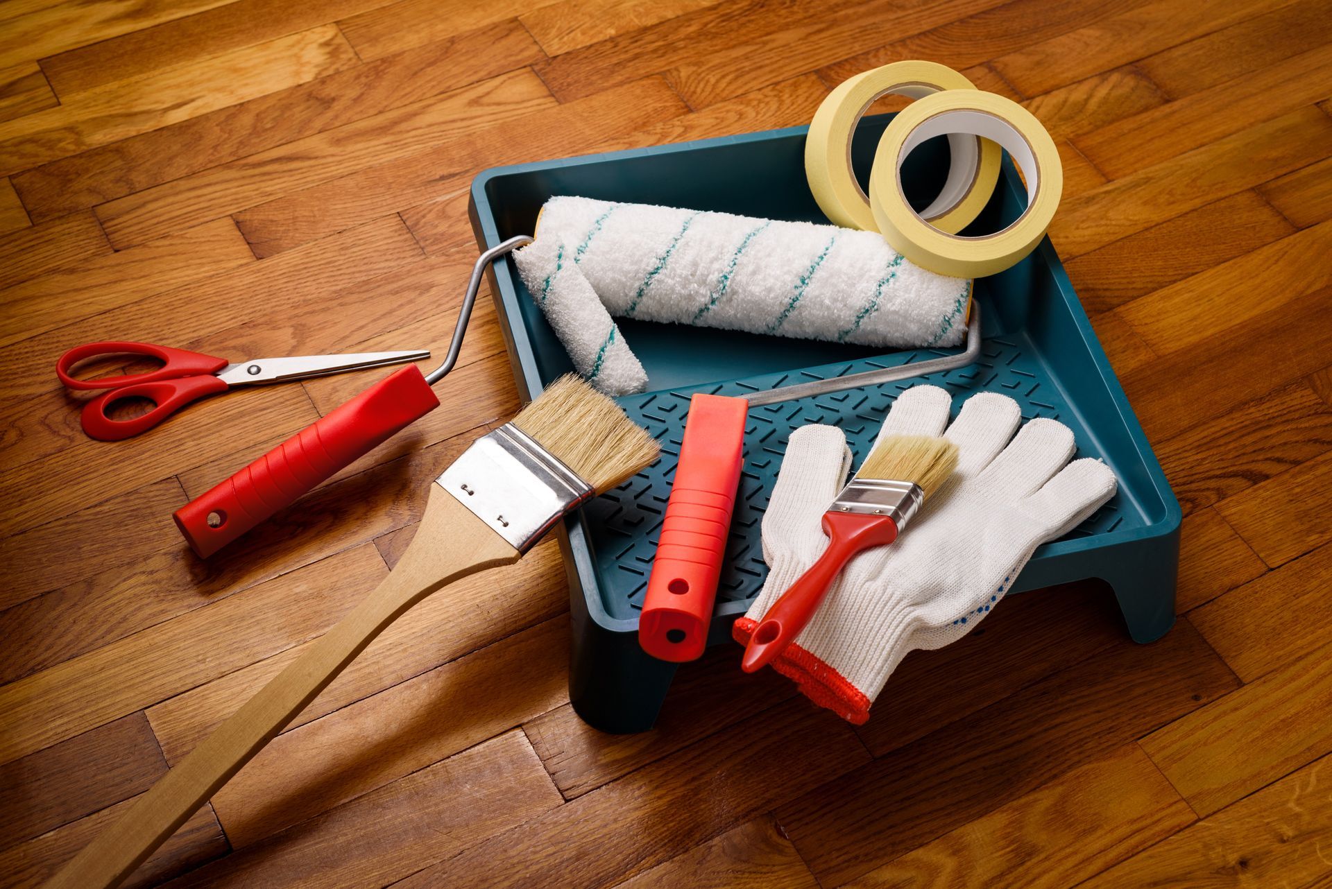Rouleaux à peinture, pinceaux, rubans adhésifs, ciseaux et gants dans un bac bleu sur un sol en bois