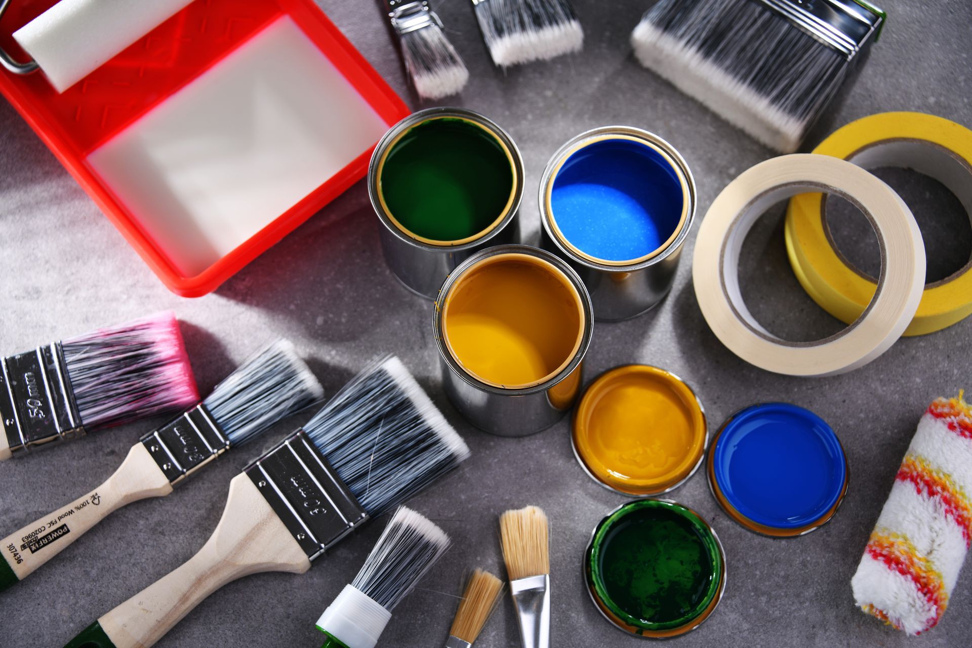 Matériel de peinture comprenant des pinceaux, des pots de peinture, des rouleaux, des rubans adhésifs et un bac rouge sur une surface grise.