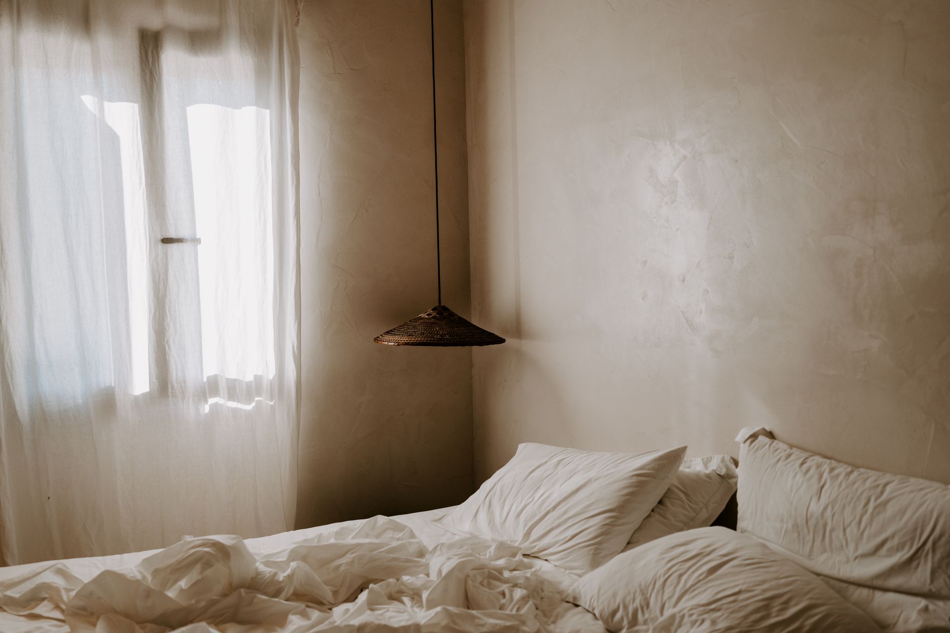 Chambre beige clair avec linge de lit blanc et une fenêtre recouverte de voilages.