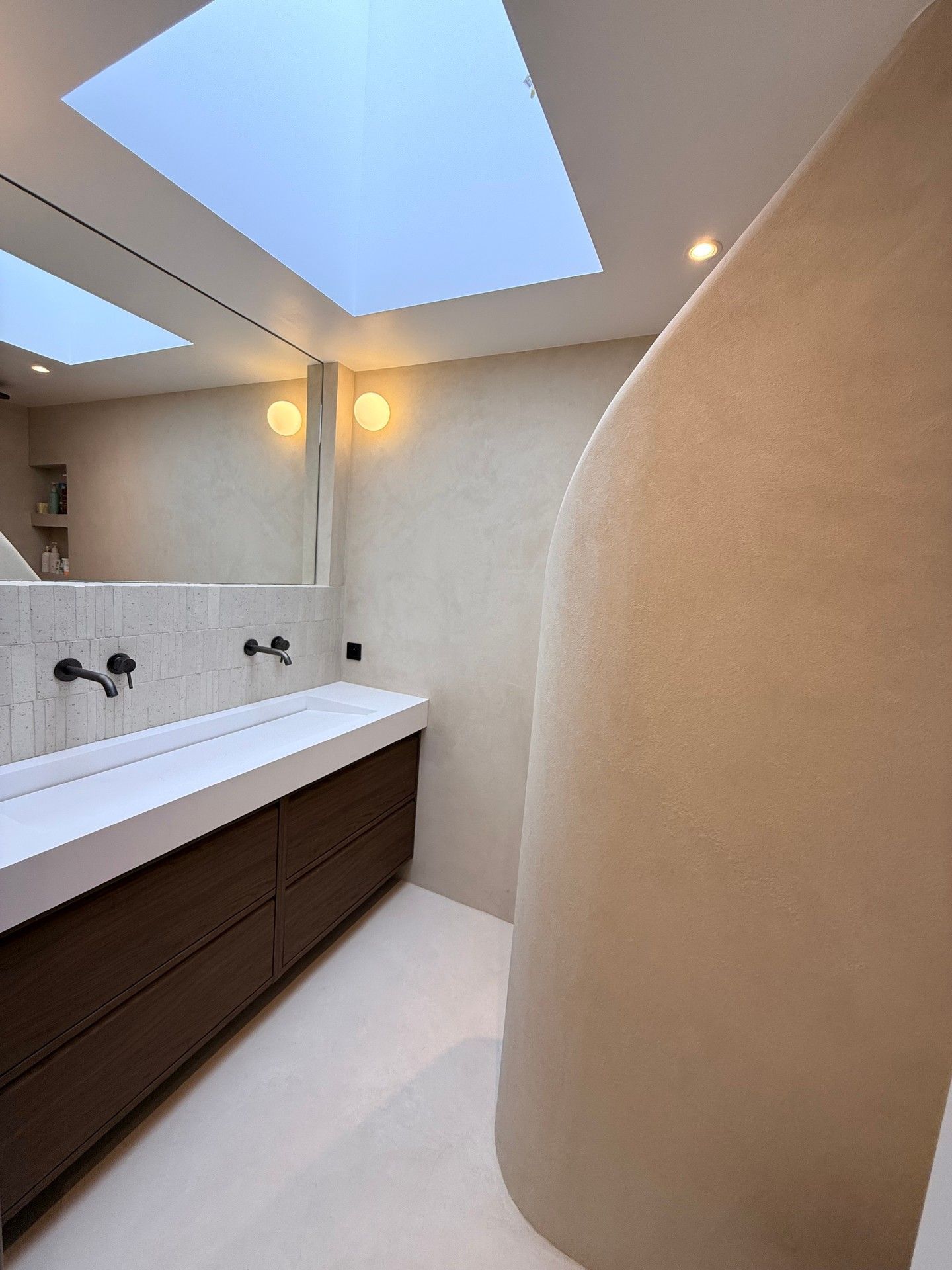 Salle de bains moderne avec puits de lumière, long meuble lavabo blanc, armoires en bois et mur beige incurvé.