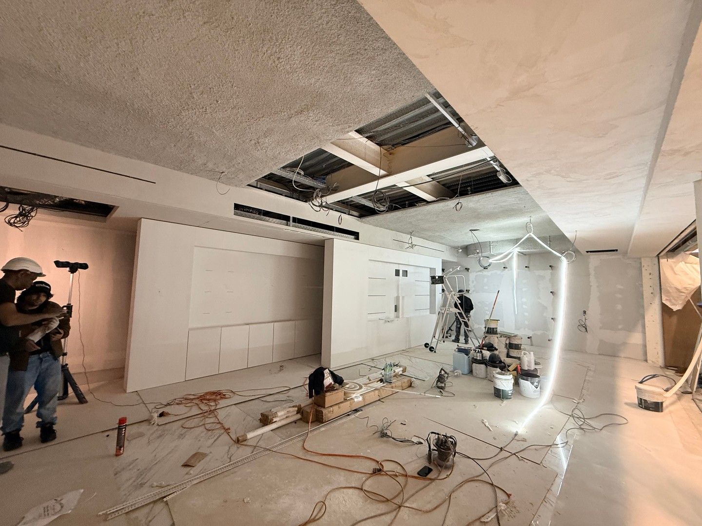 Travaux de rénovation dans une pièce, panneaux de plafond retirés, outils et câbles au sol, ouvriers présents.
