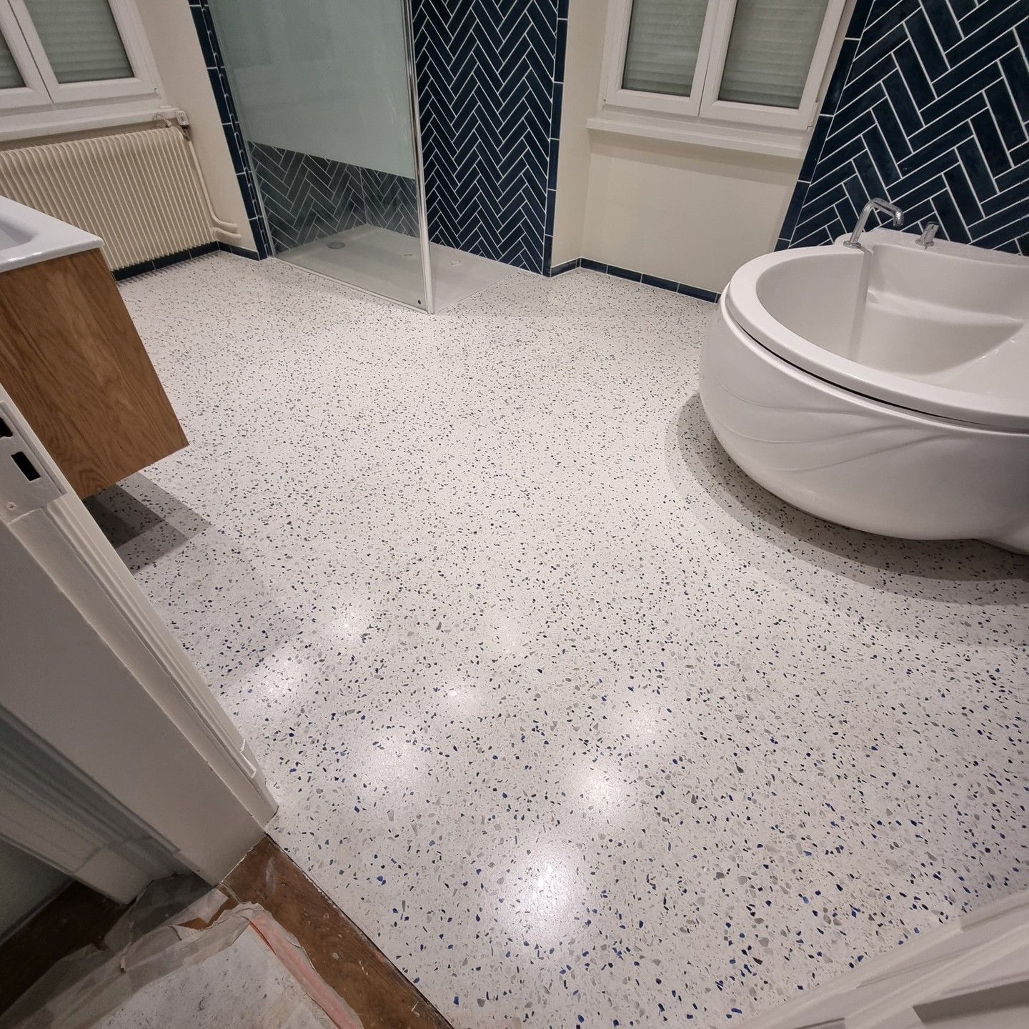 Salle de bains avec sol en terrazzo, porte de douche en verre et baignoire blanche près d'une fenêtre.