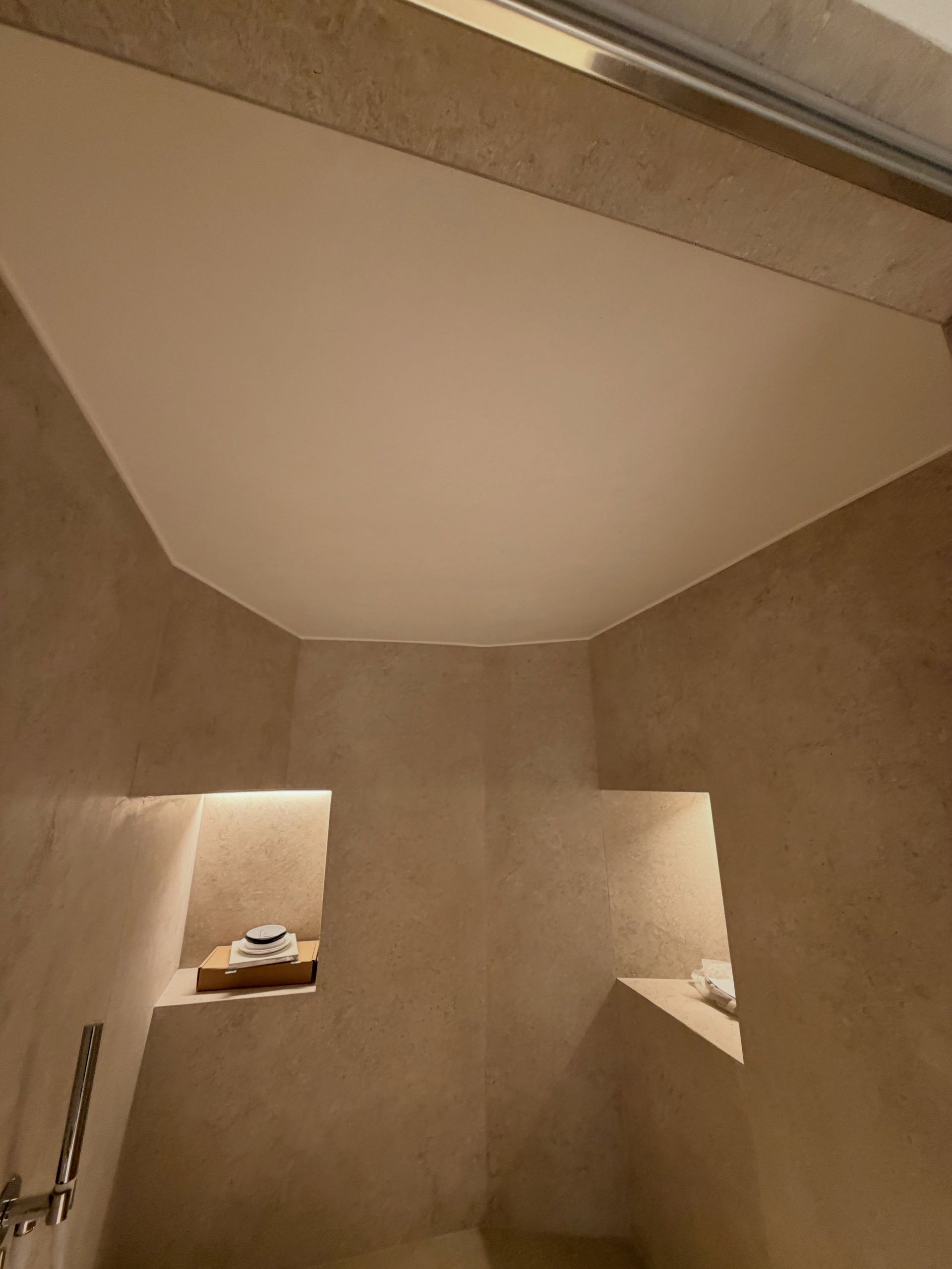 Petite douche carrelée beige avec un plafond haut et incliné et des niches intégrées, dont une pour ranger les serviettes pliées.