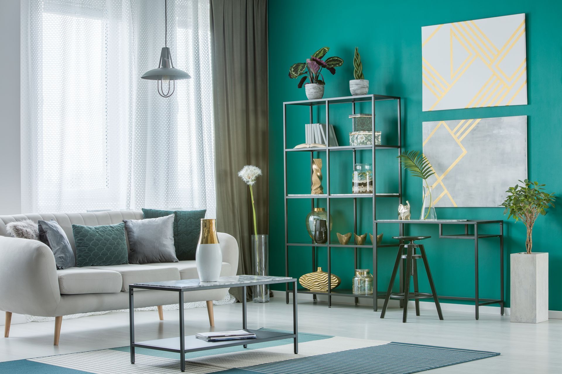 Salon bleu sarcelle lumineux avec canapé gris, table basse en verre, étagères et tableaux muraux abstraits.