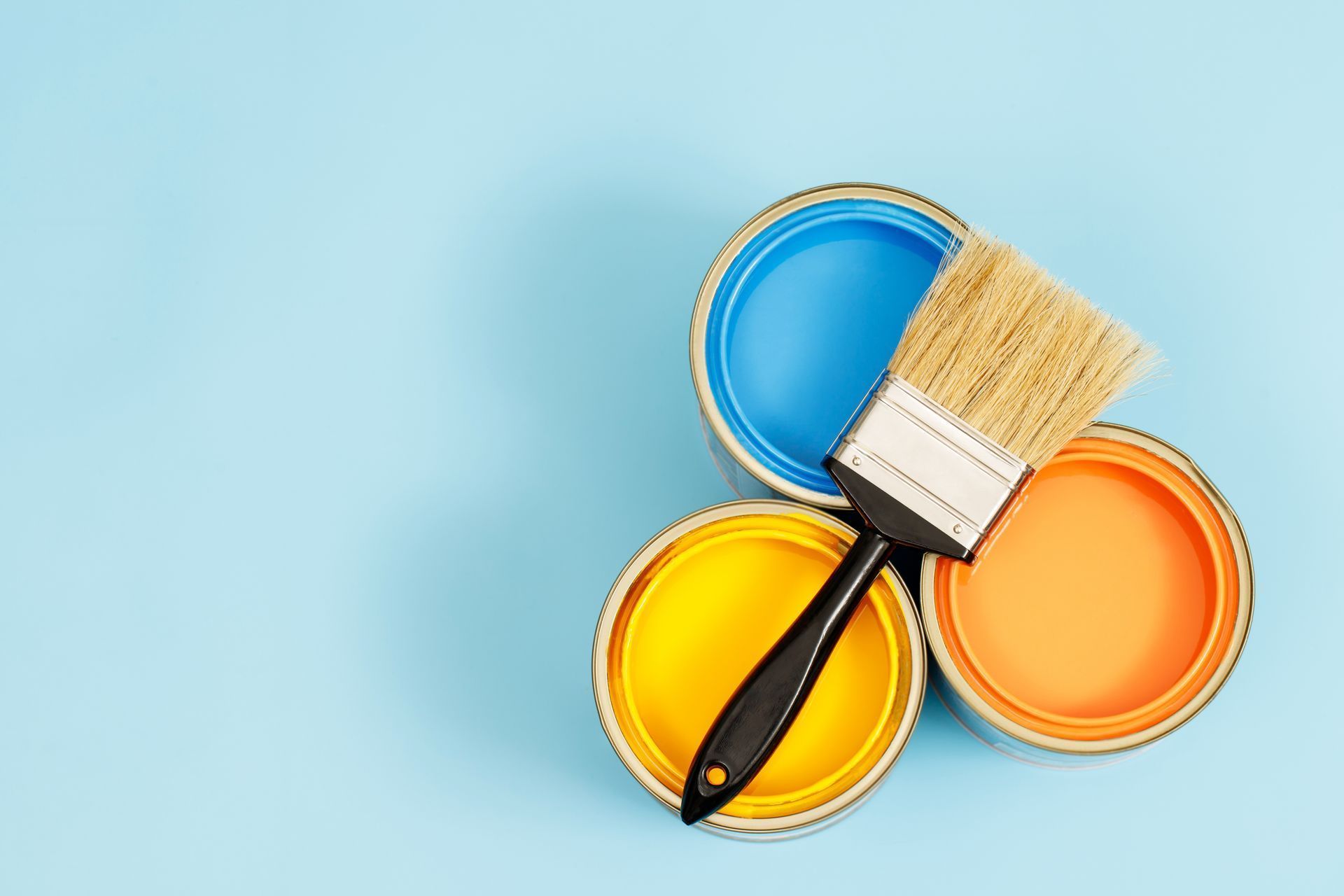 Un pinceau posé sur trois pots de peinture ouverts, bleu, jaune et orange, sur un fond bleu clair.