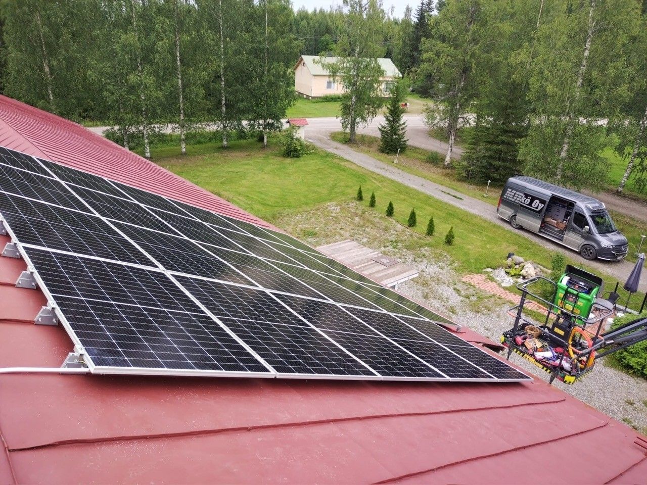 Aurinkopaneelit punaisella katolla ajotien vieressä, taustalla pakettiauto ja puita.