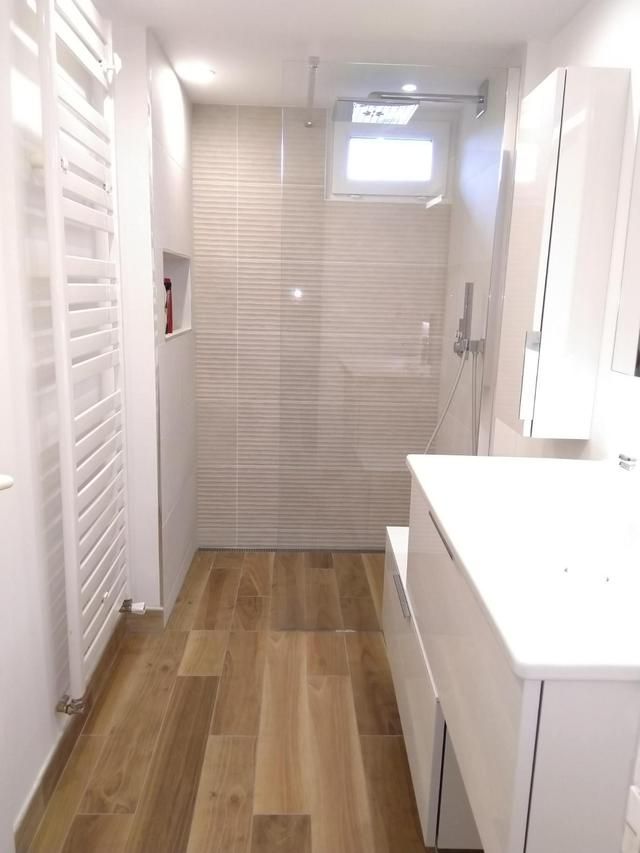 Une salle de bains étroite et moderne avec un sol imitation bois, une douche en verre, un lavabo et un meuble-lavabo blancs.