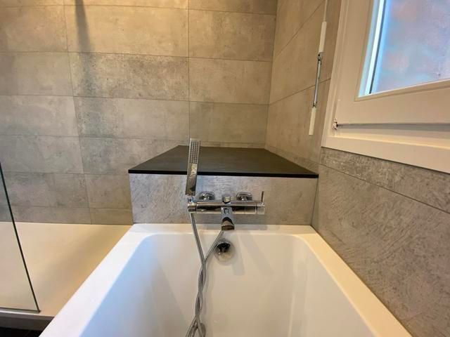 Baignoire avec robinet et pommeau de douche, entourée de murs carrelés gris et d'une étagère noire.