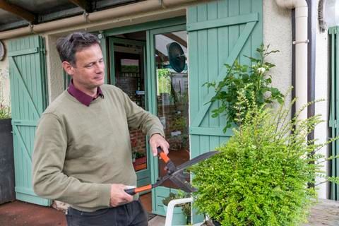 Un homme taille un buisson vert avec des cisailles devant un bâtiment aux volets verts.