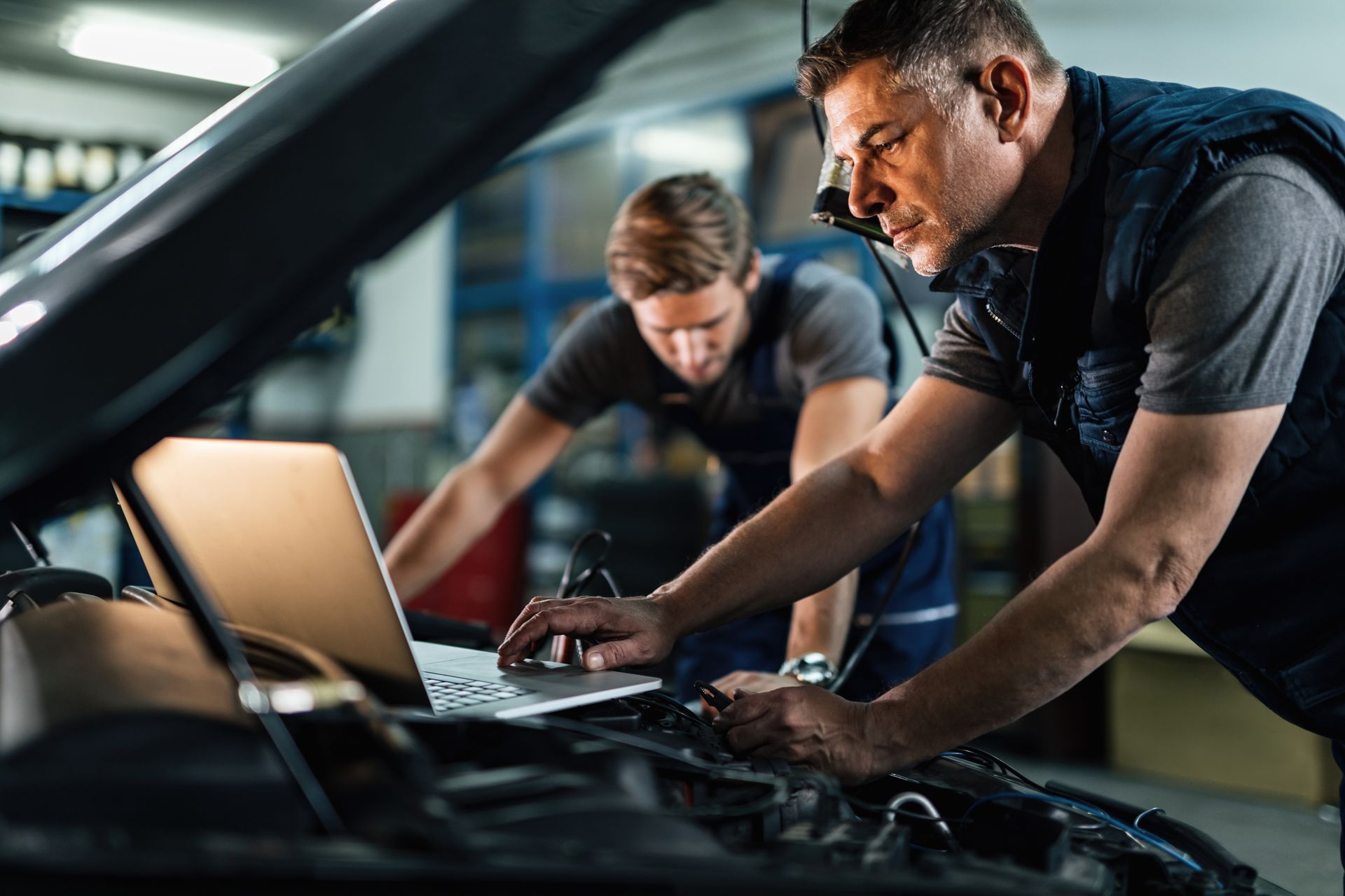 Deux mécaniciens travaillent sous le capot d’une voiture avec un ordinateur portable dans un garage