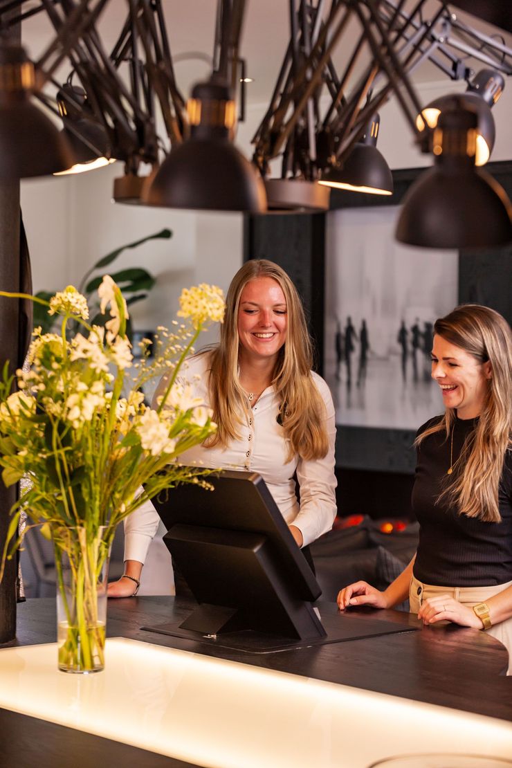 Twee vrouwen glimlachen bij een toonbank met een beeldscherm, bloemen en plafondlampen.