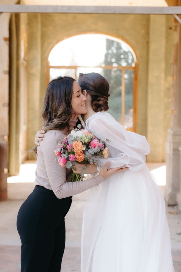 Una mujer con pantalones negros abraza a una novia con vestido blanco, sosteniendo un ramo. Afuera, arcos al fondo.