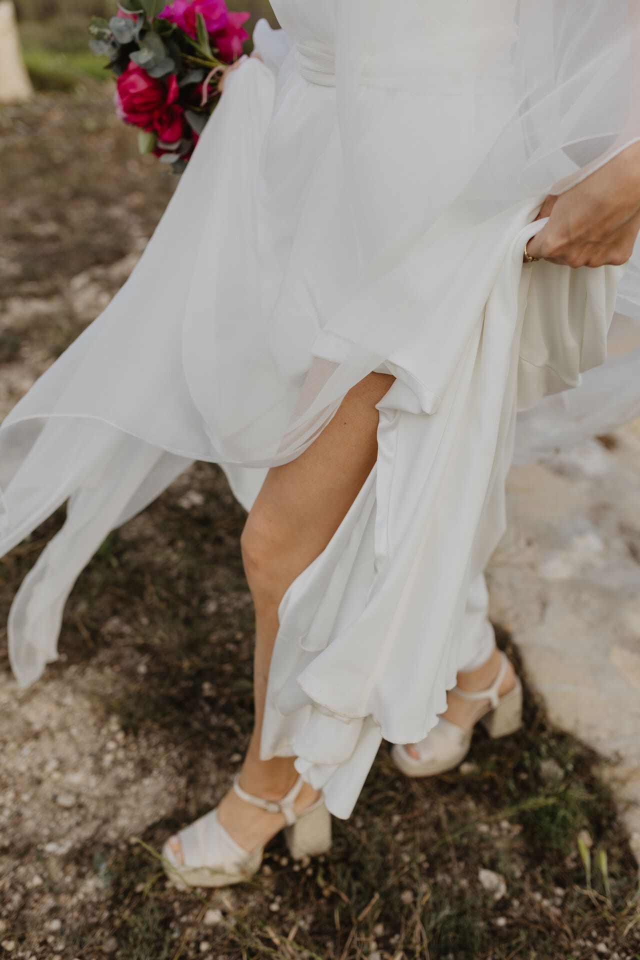 Novia con vestido blanco con abertura en la pierna, sandalias de plataforma y sosteniendo un ramo, caminando sobre suelo rocoso.