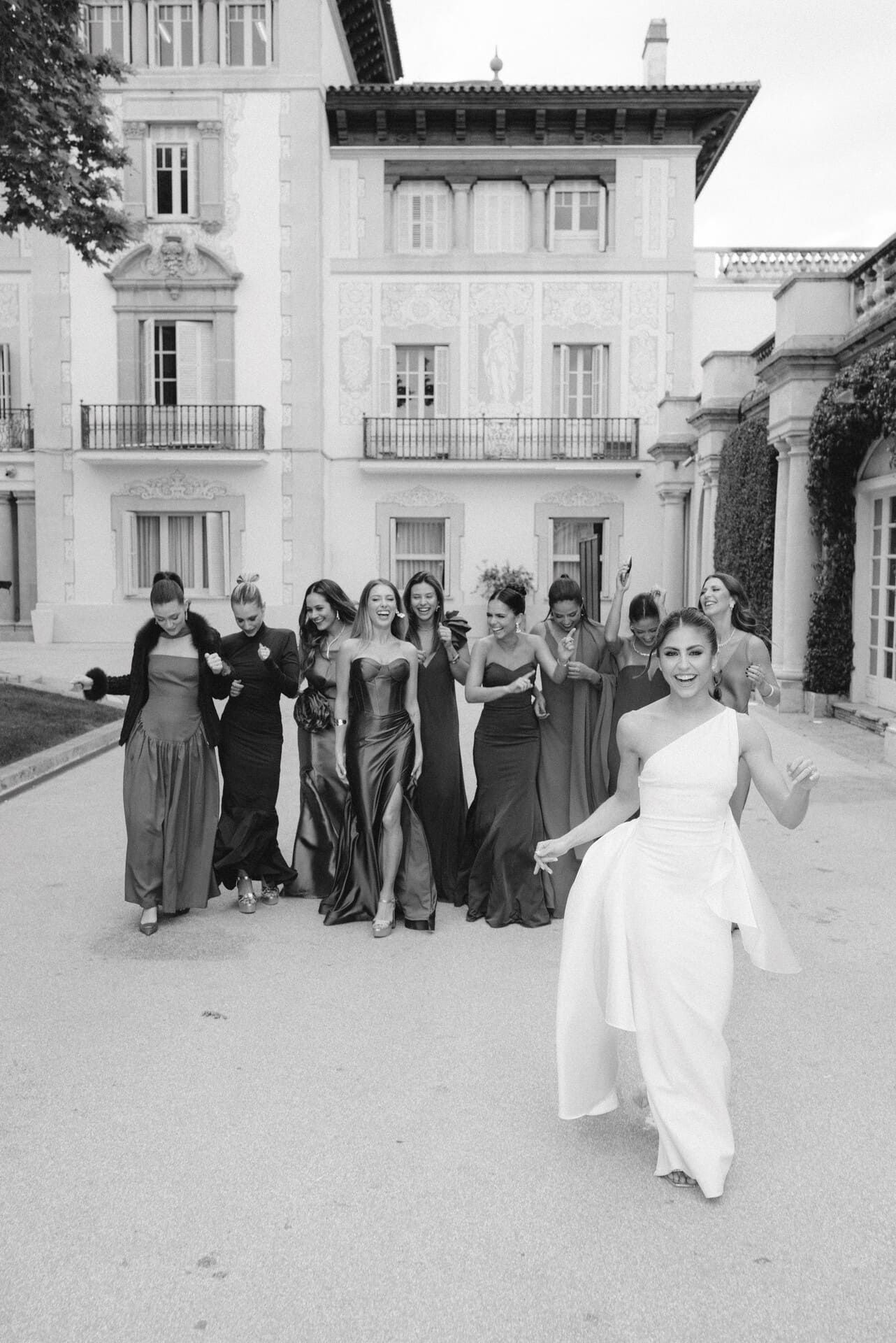 Un grupo de mujeres con vestidos posa al aire libre, con un edificio ornamentado de fondo. Una mujer con vestido blanco avanza.