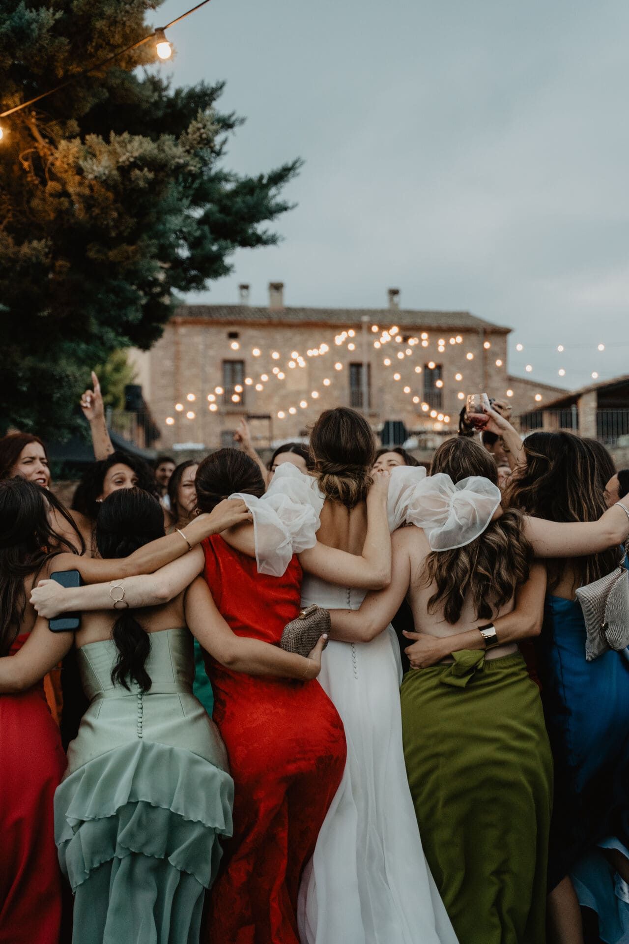 Las damas de honor con vestidos coloridos abrazan a la novia en un patio, iluminado por luces de cadena, con un edificio al fondo.