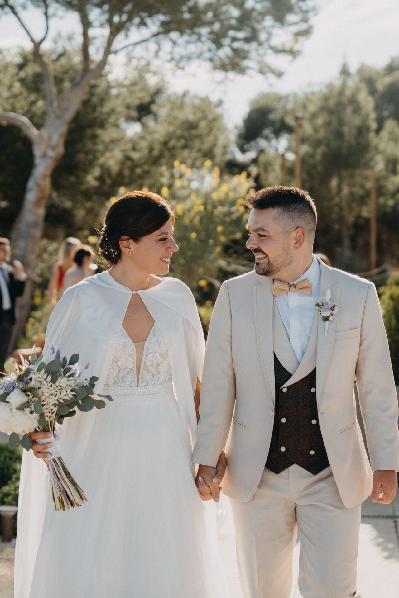 Los novios, tomados de la mano y sonriéndose, caminan al aire libre. Ella lleva un vestido blanco y él un traje color canela.