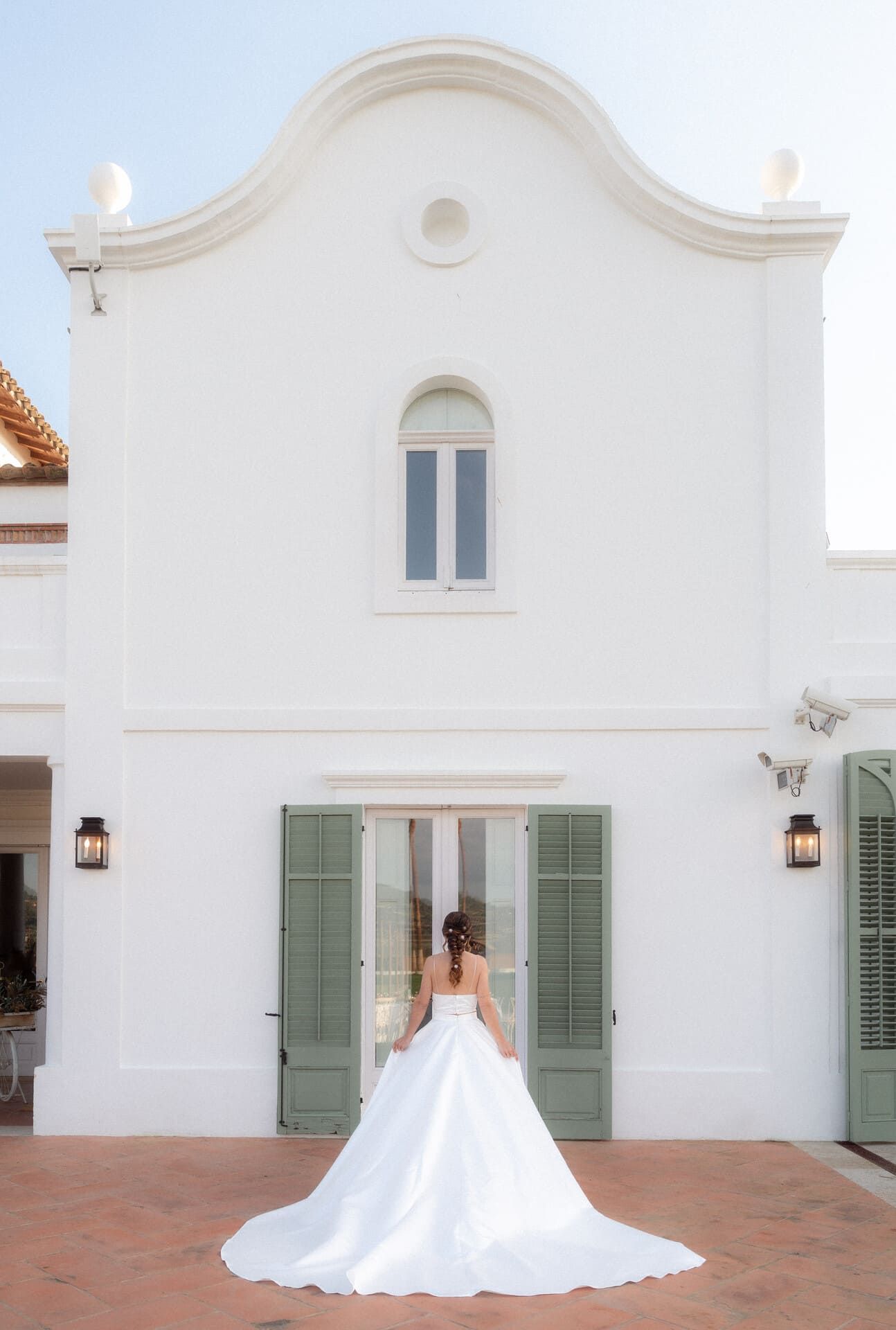 Una novia con un vestido blanco se encuentra frente a un edificio blanco con contraventanas verdes.