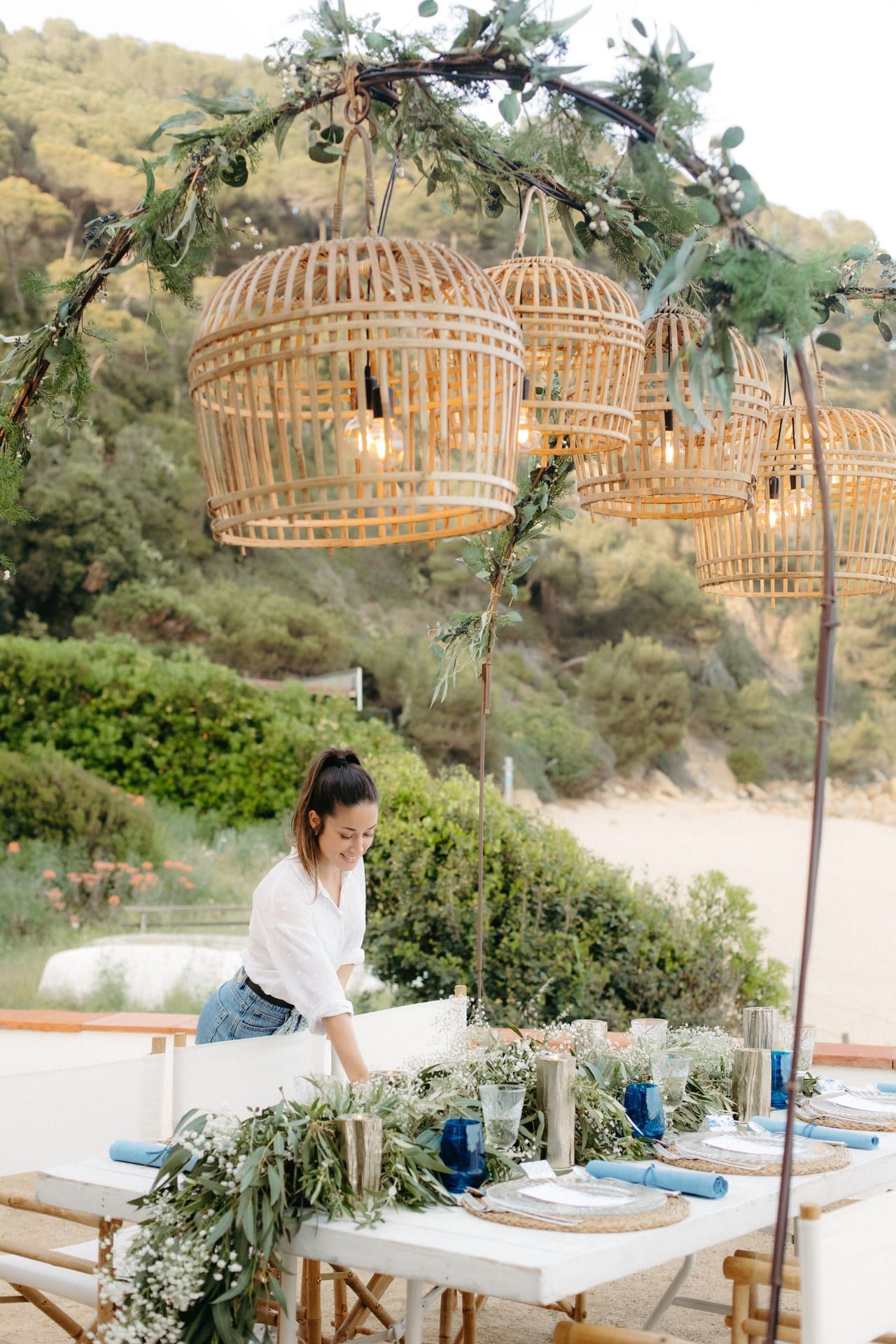 Mujer colocando una mesa con detalles azules bajo luces colgantes tejidas en un entorno de playa.