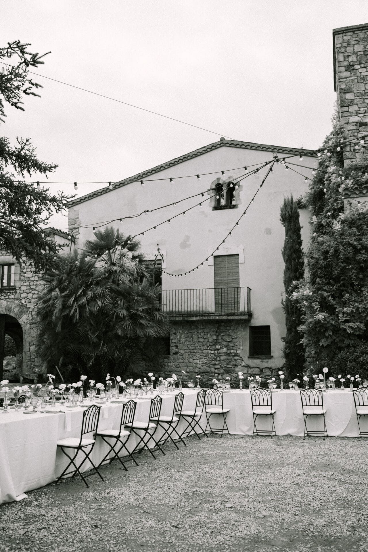 Configuración de recepción de boda al aire libre en blanco y negro con mesas largas, sillas y un edificio al fondo.