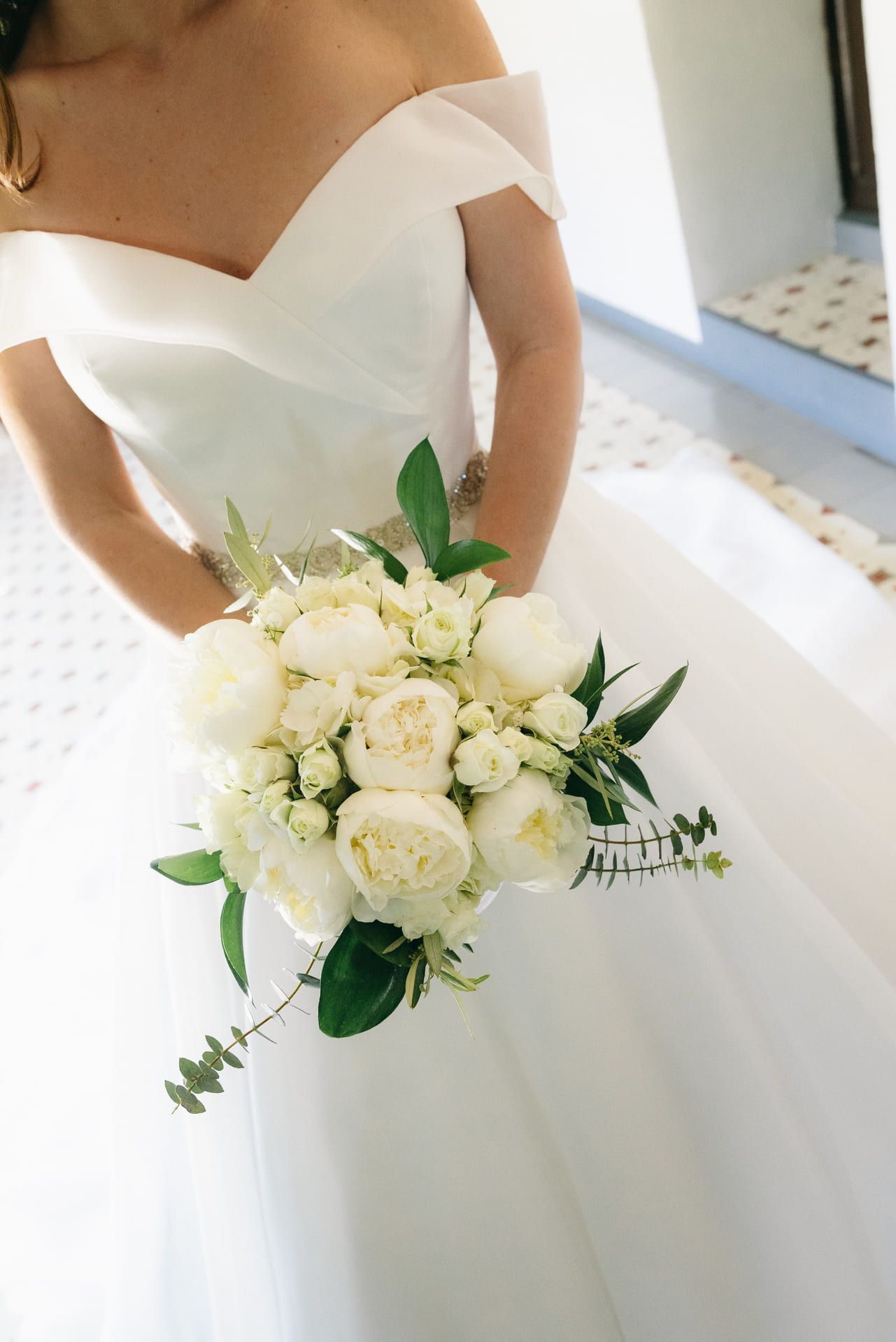 La novia con un vestido blanco con hombros descubiertos sostiene un ramo de flores blancas.