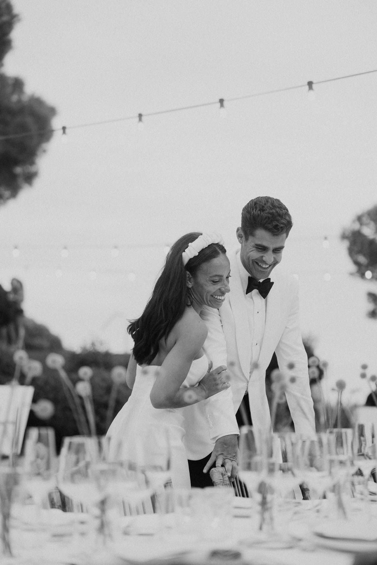 Fotografía en blanco y negro: pareja sonriente en una recepción de boda al aire libre, de pie cerca de una mesa preparada con vasos.