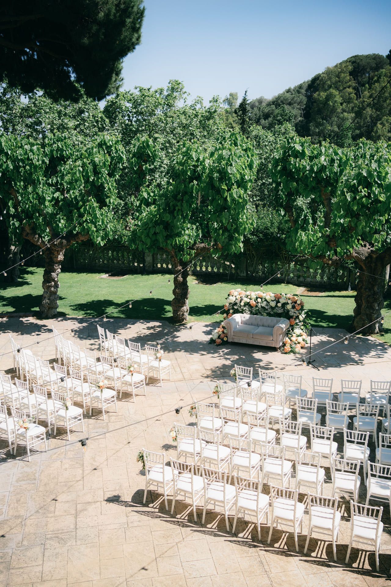 Ceremonia de boda organizada al aire libre con sillas blancas, árboles y un sofá adornado con flores como telón de fondo.