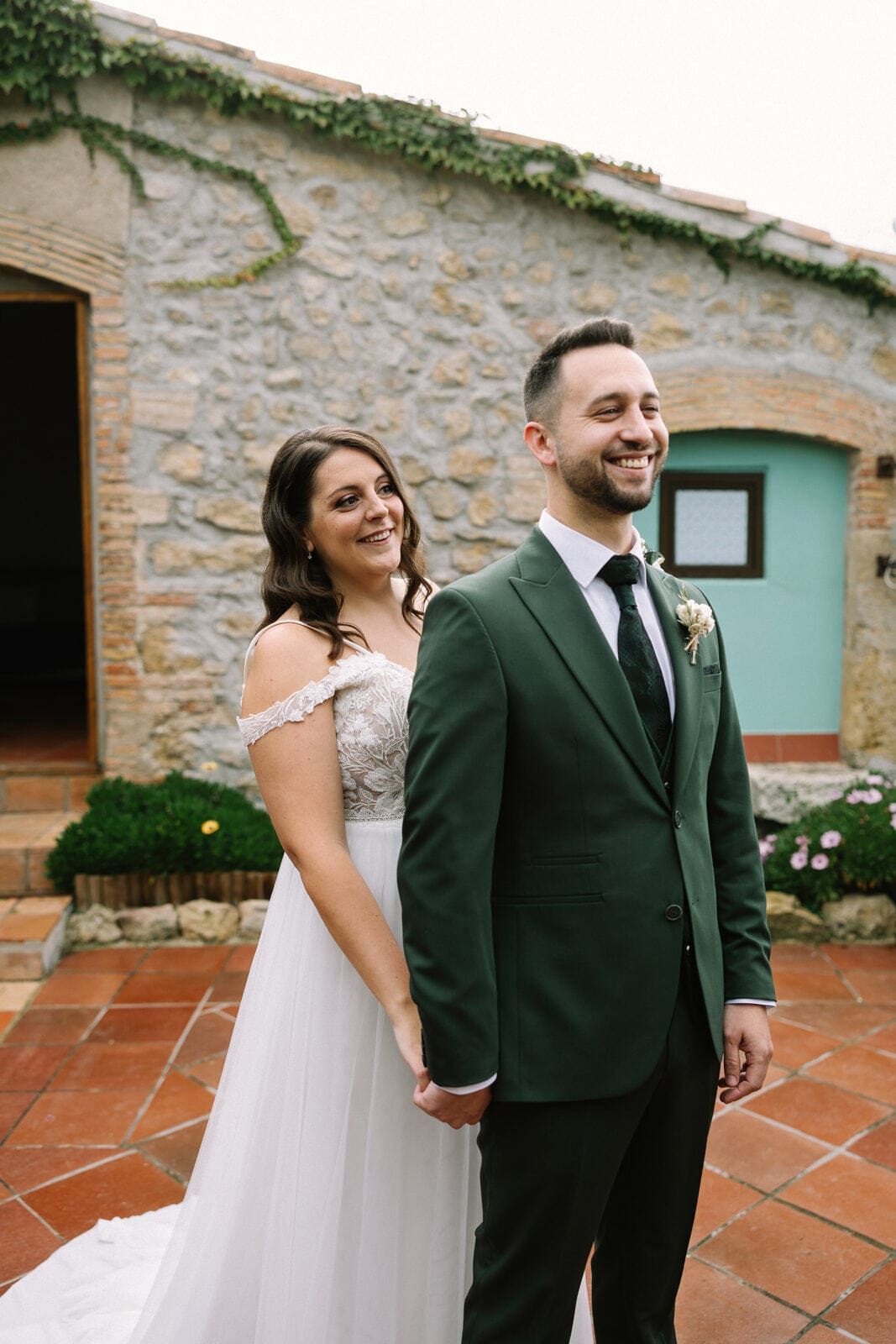 La novia, vestida de blanco, mira al novio, vestido de verde; tomados de la mano, sonriendo. Patio de ladrillos, con un edificio de piedra al fondo.