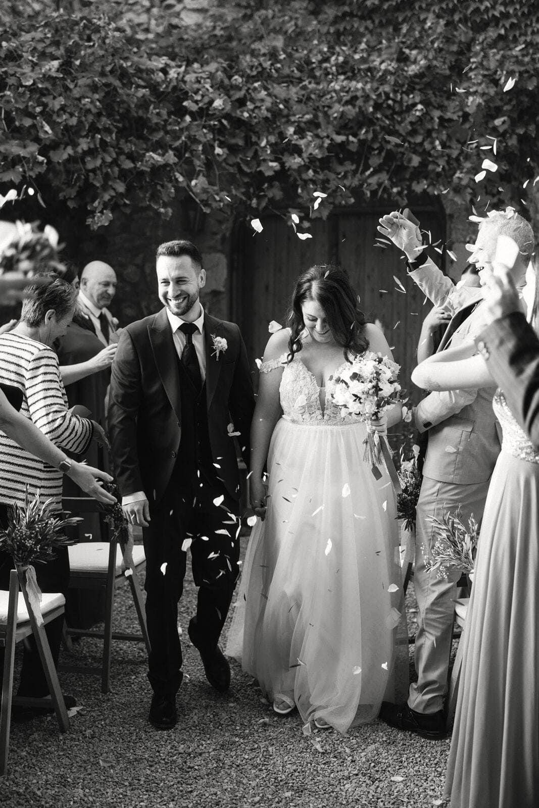 Los recién casados ​​caminan, bañados en confeti, rodeados de invitados en un jardín. Blanco y negro.