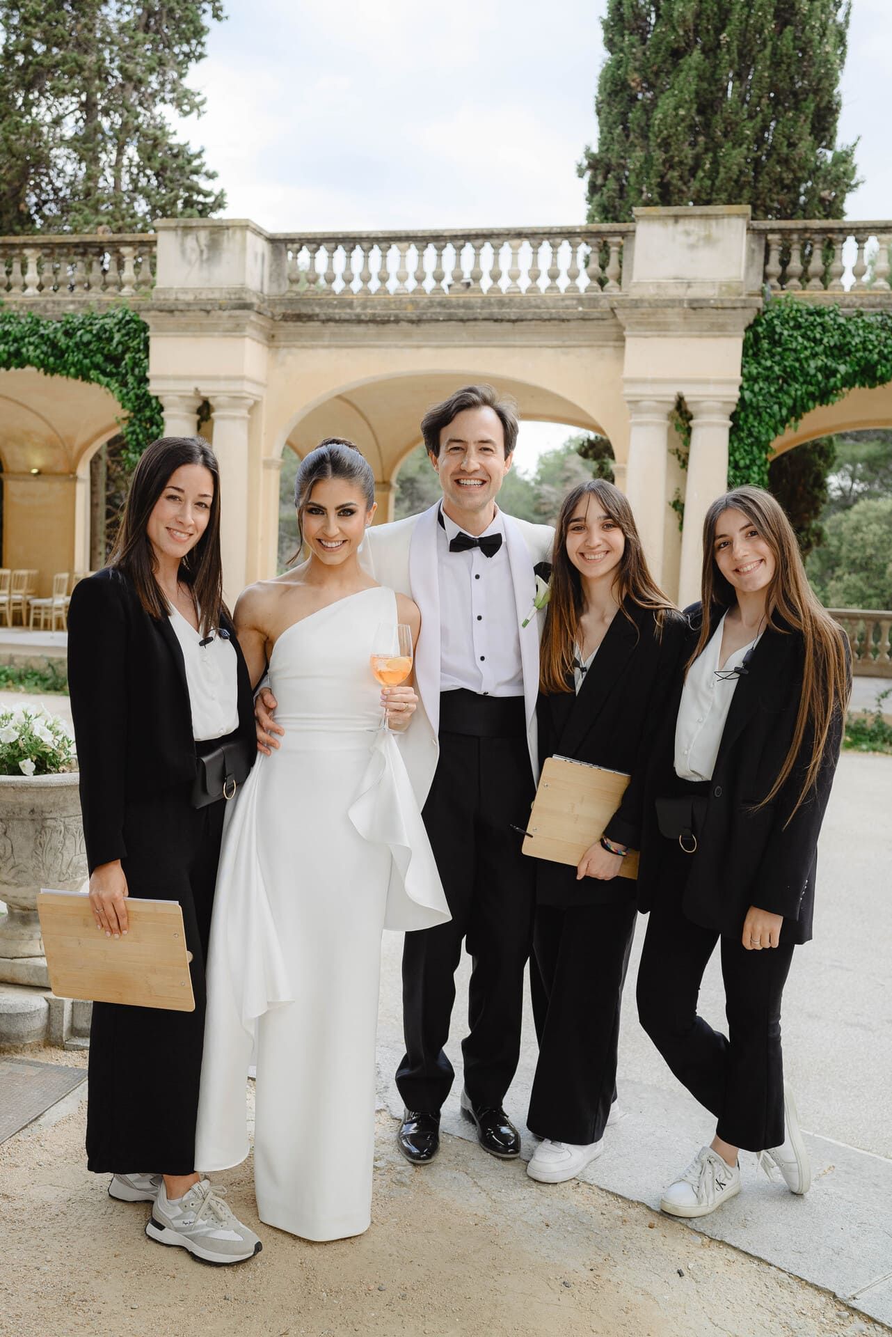 Un grupo de novios posa frente a un edificio con arcos. Una mujer con vestido blanco, un hombre con esmoquin y otros cuatro con blazers negros, todos sonriendo.