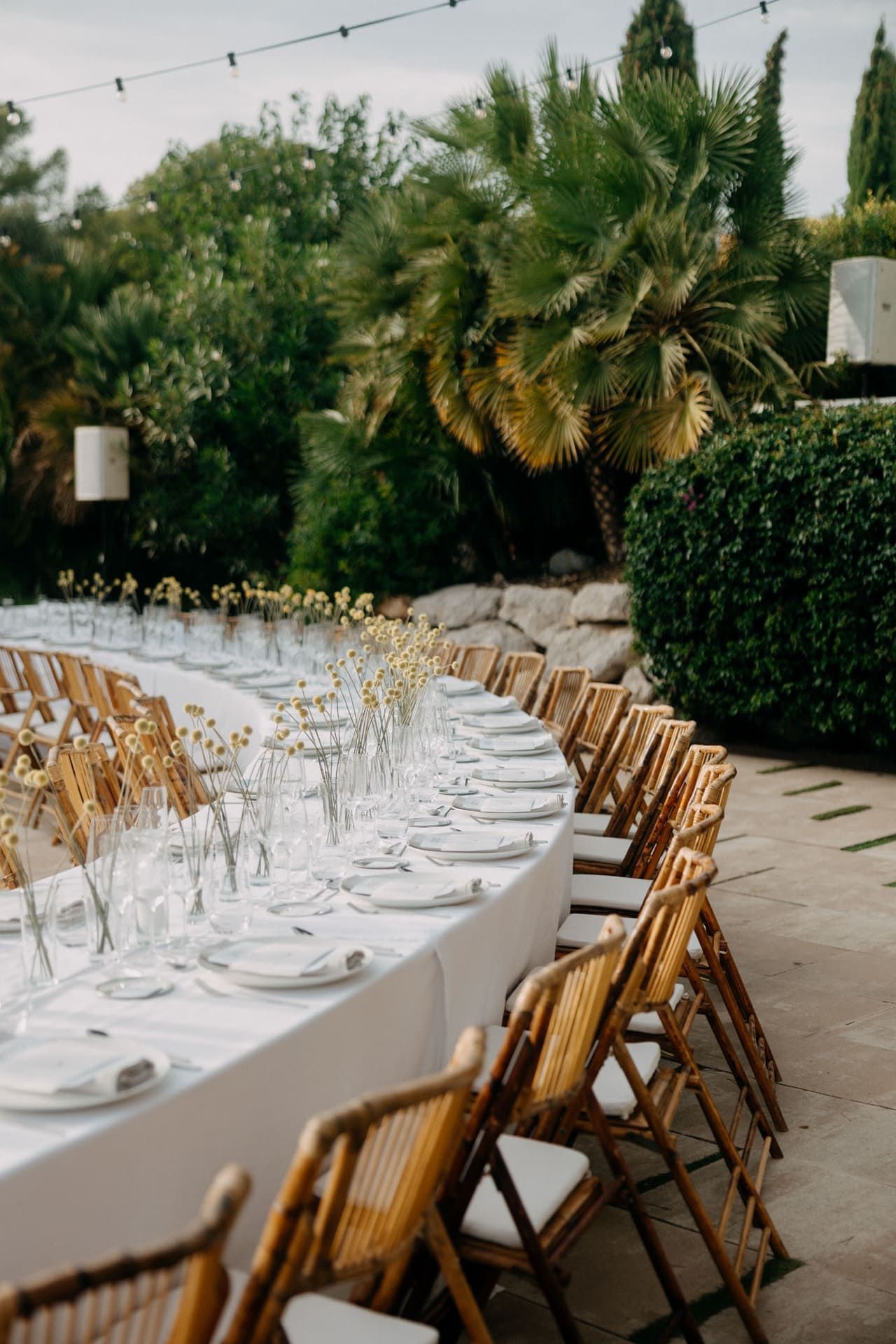 Mesa de comedor exterior larga y curva para boda, con sillas de bambú y centros de mesa florales.