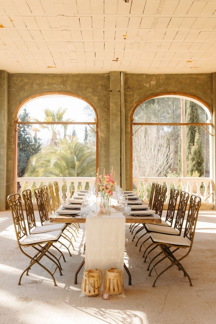 Mesa de comedor larga dispuesta para comer, en un espacio exterior iluminado por el sol y con ventanas arqueadas.