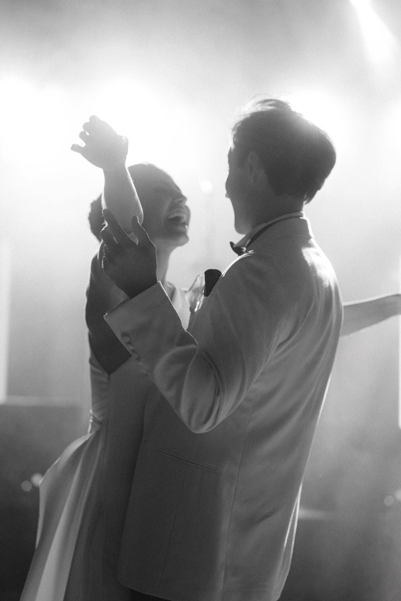 Una pareja de recién casados ​​baila, bañada por la luz; el novio sostiene un micrófono.