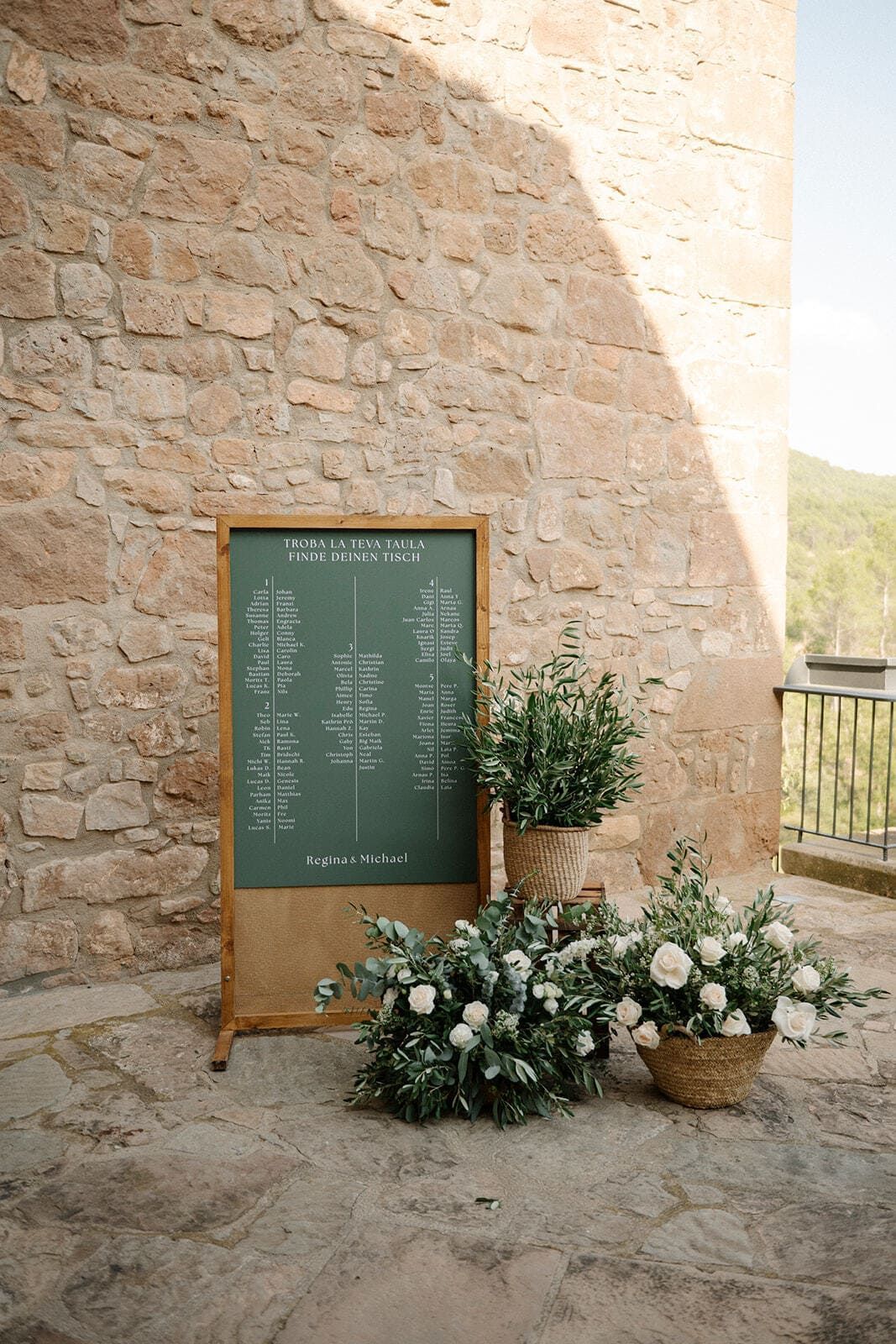 Plano de asientos para boda con vegetación, flores y un fondo rústico de pared de piedra.