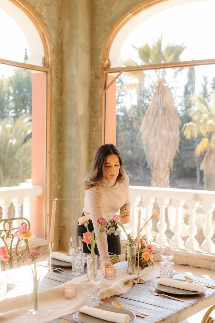 Mujer arreglando flores en una mesa preparada para cenar, al aire libre en un balcón con palmeras.