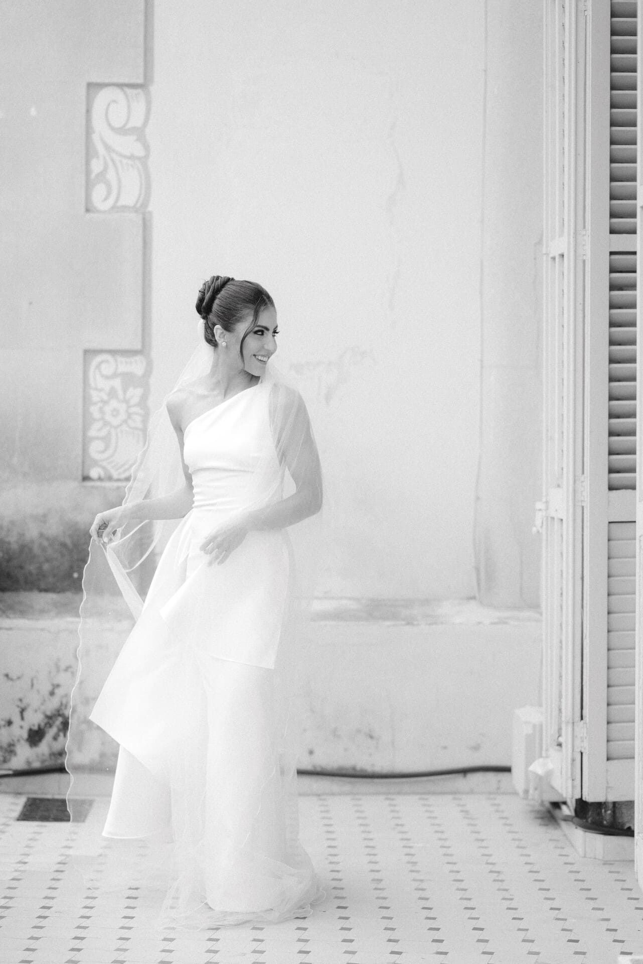 La novia con vestido blanco sonríe, dando vueltas cerca de un edificio con detalles arquitectónicos.