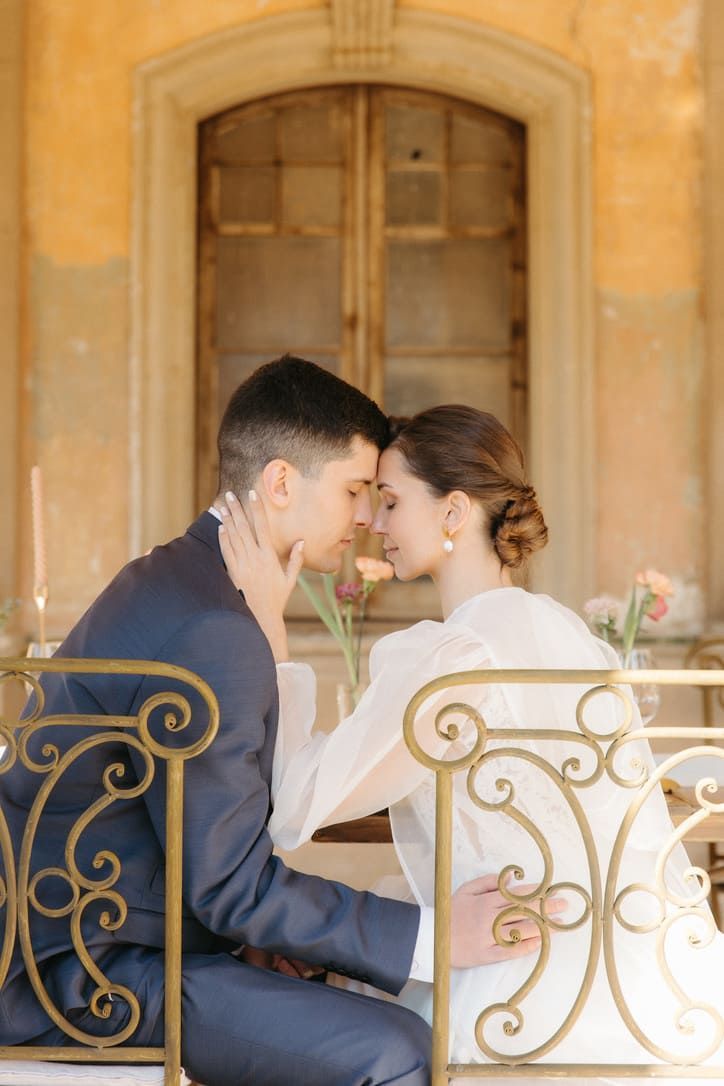 Pareja tocándose la frente, sentada en una mesa en un balcón, con una ventana al fondo.