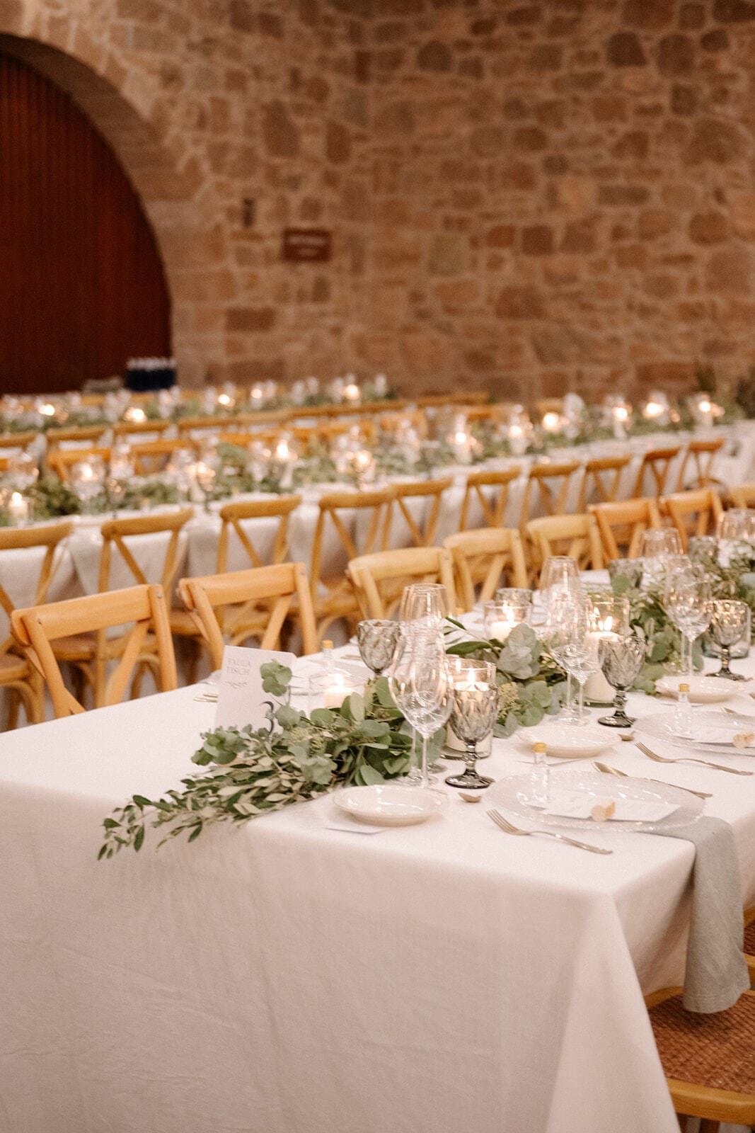 Mesas largas preparadas para una recepción de boda con velas, vegetación y elegantes cubiertos en una sala con paredes de piedra.