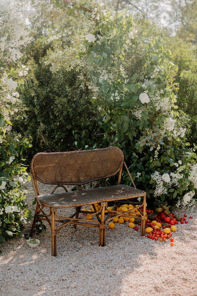 Banco de mimbre en un jardín, rodeado de flores blancas y limones y tomates esparcidos.