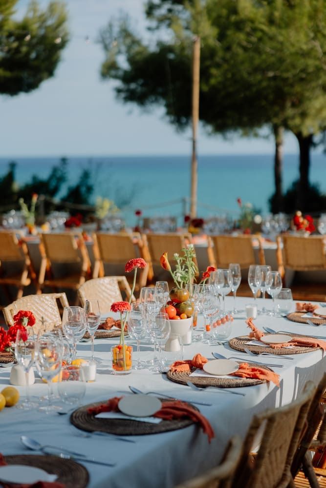 Mesa de comedor exterior para boda con fondo marino. Flores rojas y detalles en naranja.