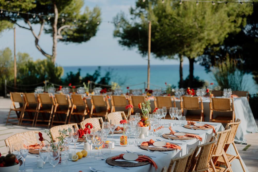 Recepción de boda al aire libre con mesas largas con vista al océano, centros de mesa florales y sillas de madera.
