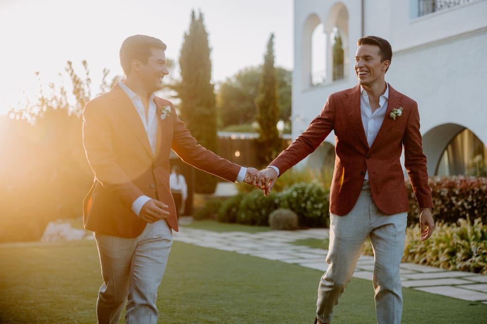 Dos hombres trajeados, tomados de la mano y sonriendo al aire libre. El atardecer proyecta un cálido resplandor.