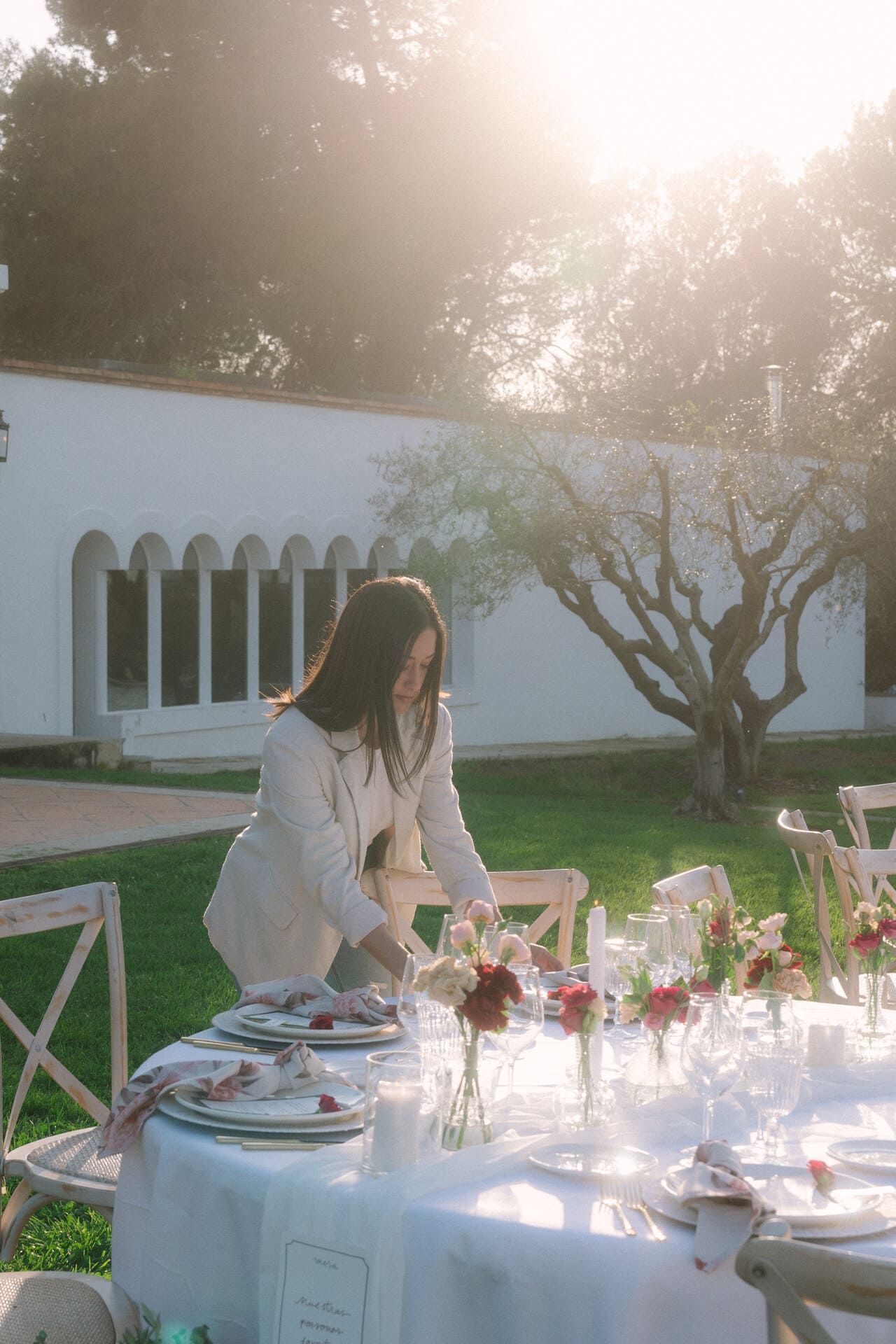 Mujer poniendo una mesa al aire libre; mantel blanco, centros de mesa florales, iluminado por el sol.