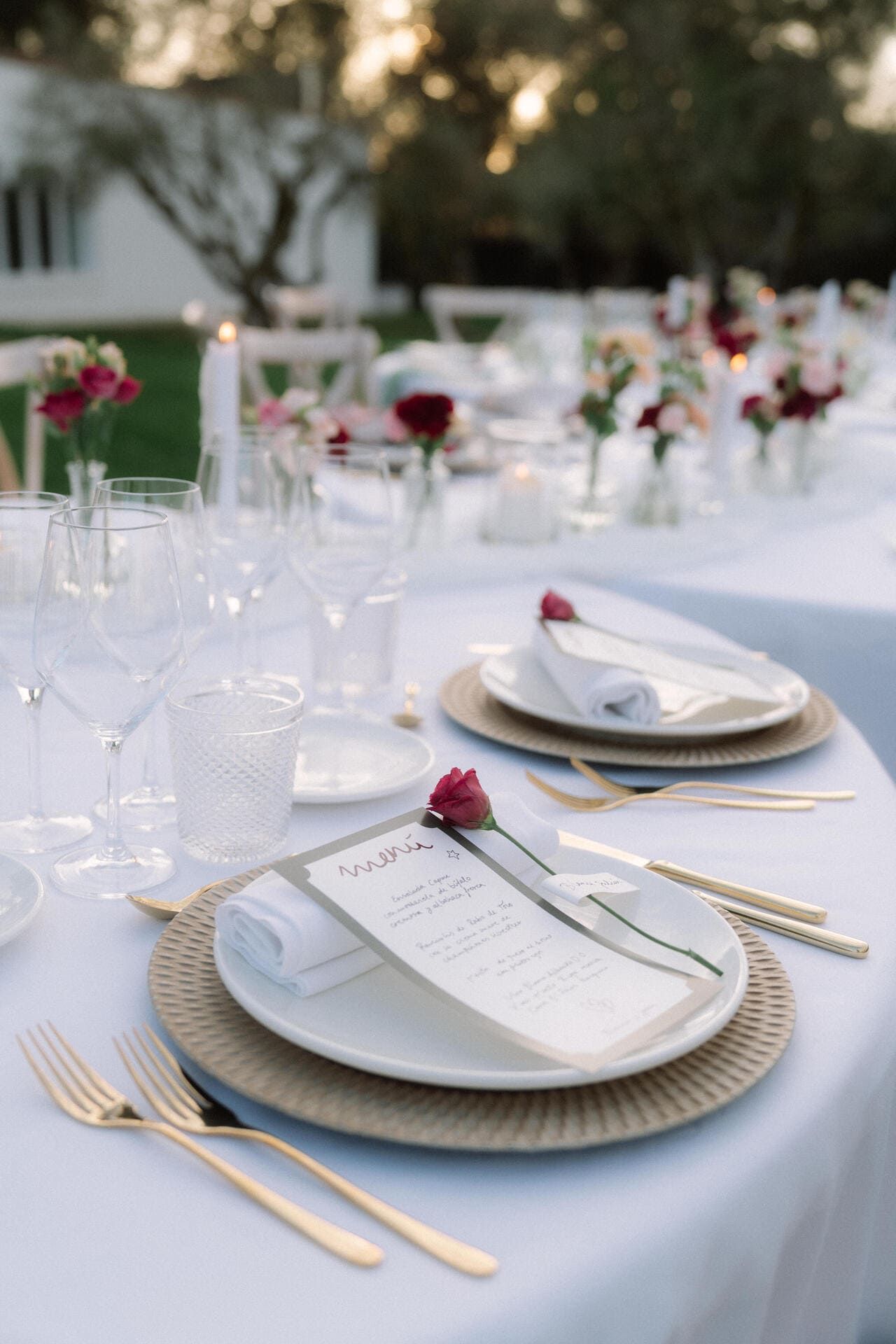 Elegante juego de mesa de comedor para exterior con mantel blanco, cubiertos dorados y centros de mesa florales.