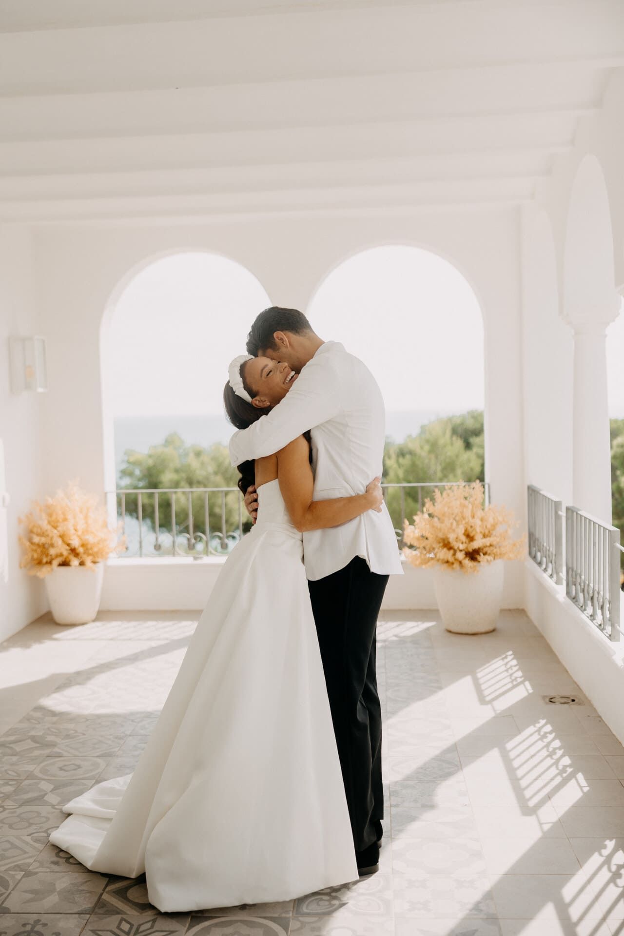 Los novios se abrazan, se besan en un balcón con arcos y vista al mar; día luminoso y soleado.