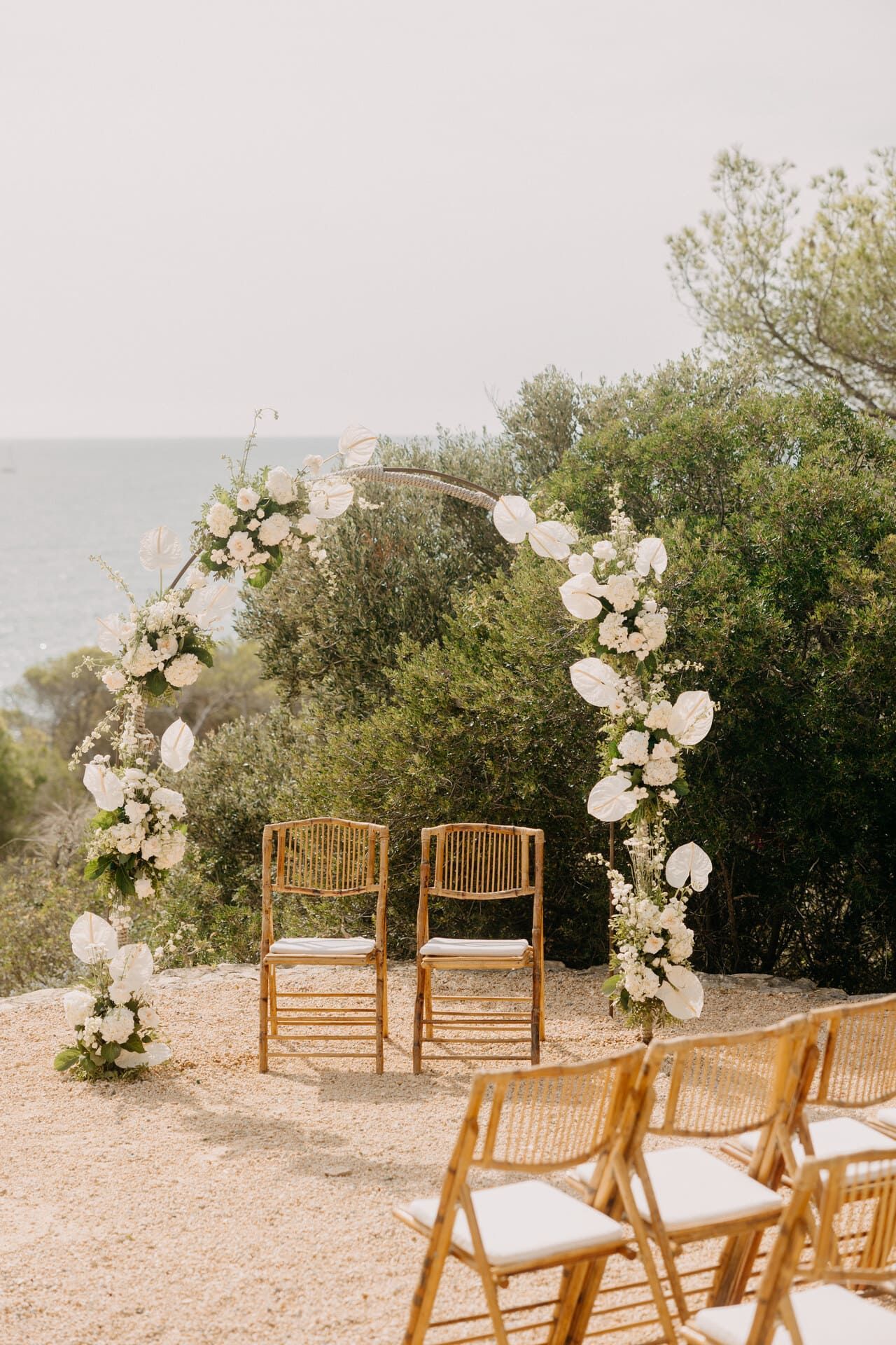 Montaje de ceremonia de boda en una ladera: arco con flores blancas, dos sillas y vista al mar.