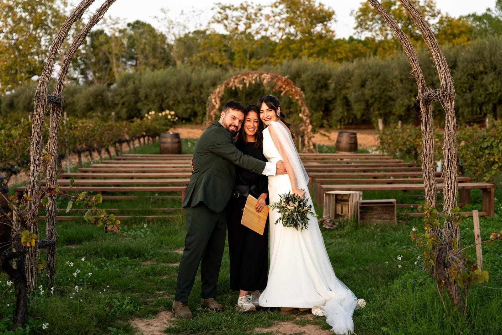 Novia y novio abrazando al oficiante bajo un arco, con un viñedo de fondo.