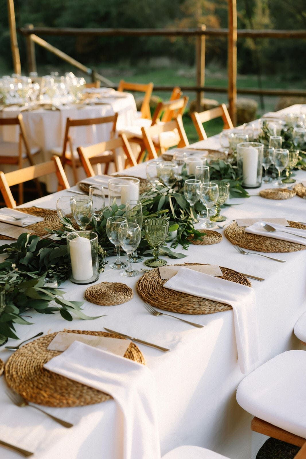 Mesa de recepción de boda al aire libre con mantelería blanca, vegetación y cubiertos.