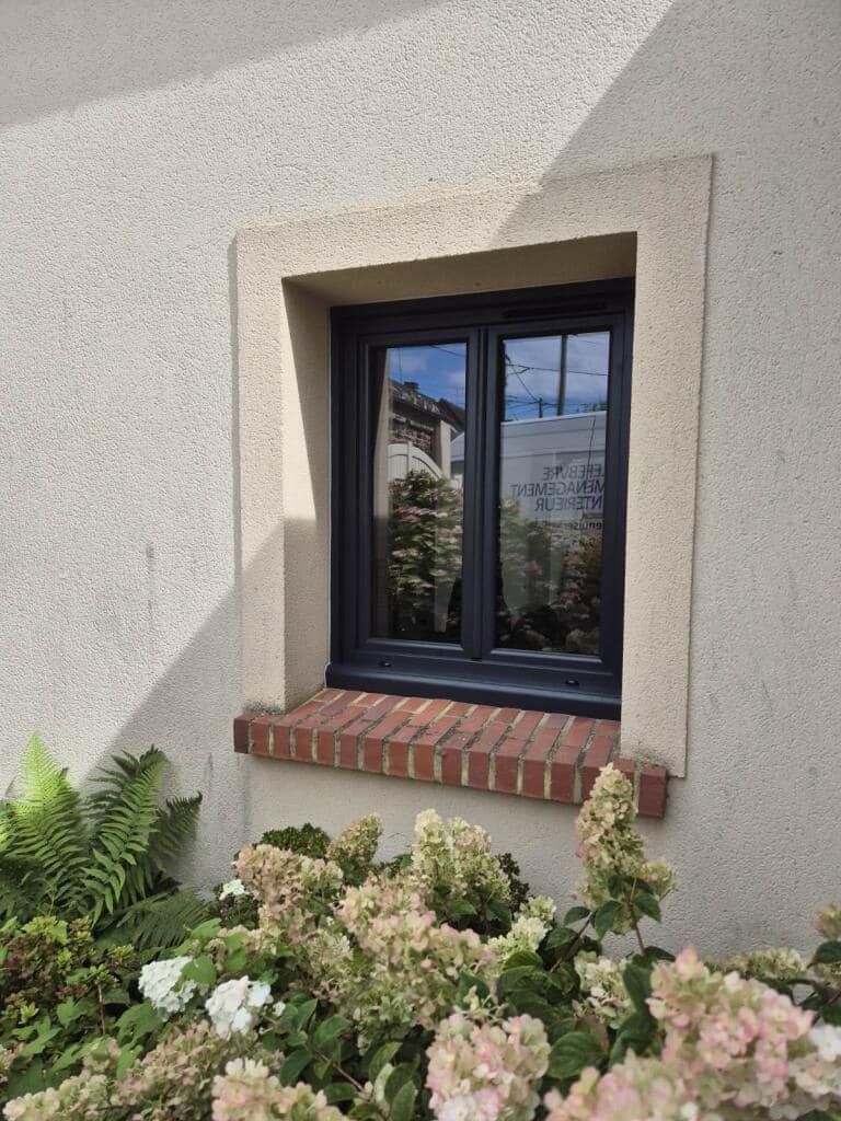 Fenêtre à cadre noir, appui de fenêtre en briques, entourée d'un mur en stuc beige.