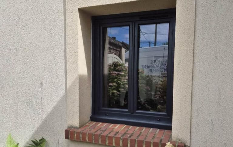 Fenêtre sombre encastrée dans un mur beige. Appui de fenêtre en briques rouges en dessous.